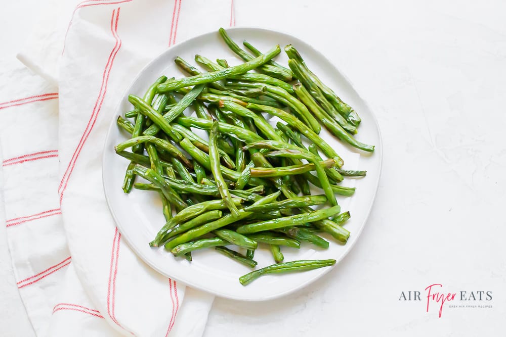 cooked green beans on a white plate next to a red striped kitchen towel on a marble countertop