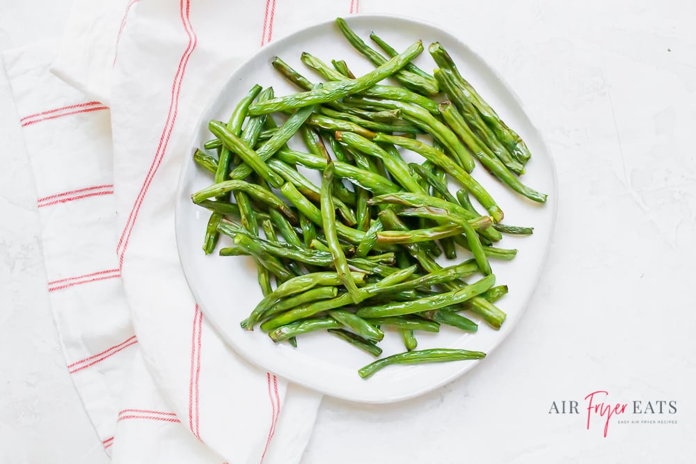 cooked green beans on a white plate next to a red striped kitchen towel on a marble countertop