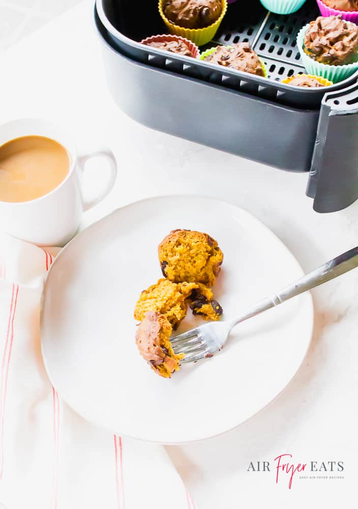 Top part of the photo is a black air fryer basket filled with pumpkin muffins in colorful muffin cups. Towards the middle is a white cup with coffee, hinted with cream. Below that is a white plate with a muffin cut in half with a fork. All set on a white background with a white/red striped napkin to the left of the plate.