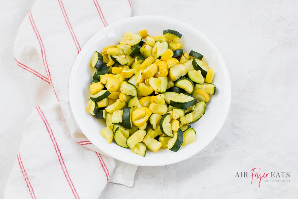 Air fryer yellow squash and zucchini cooked in a white bowl on a white back ground with a white and red napkin to the left