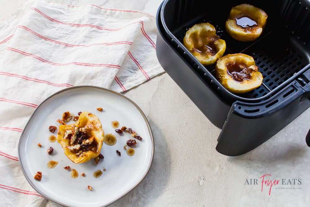 Horizontal picture with an air fryer spiced apple on a white plate on the left and apples in a black air fryer basket on the right. Then a white with a thin red stripe napkin on the left top.