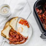 Overhead shot of coffee in a white mug to the left. Center of the photo is a white plate with air fryer bacon, toast cut into triangles, fried egg and cherry tomatoes. On the right is a black air fryer basket full of cooked bacon.