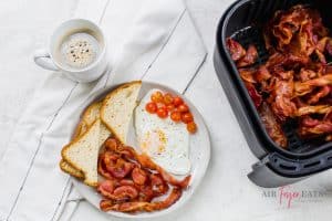Overhead shot of coffee in a white mug to the left. Center of the photo is a white plate with air fryer bacon, toast cut into triangles, fried egg and cherry tomatoes. On the right is a black air fryer basket full of cooked bacon.