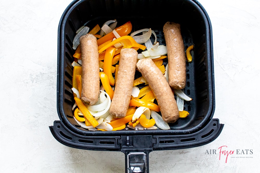 raw brats on top of cooked peppers and onions in a black air fryer basket