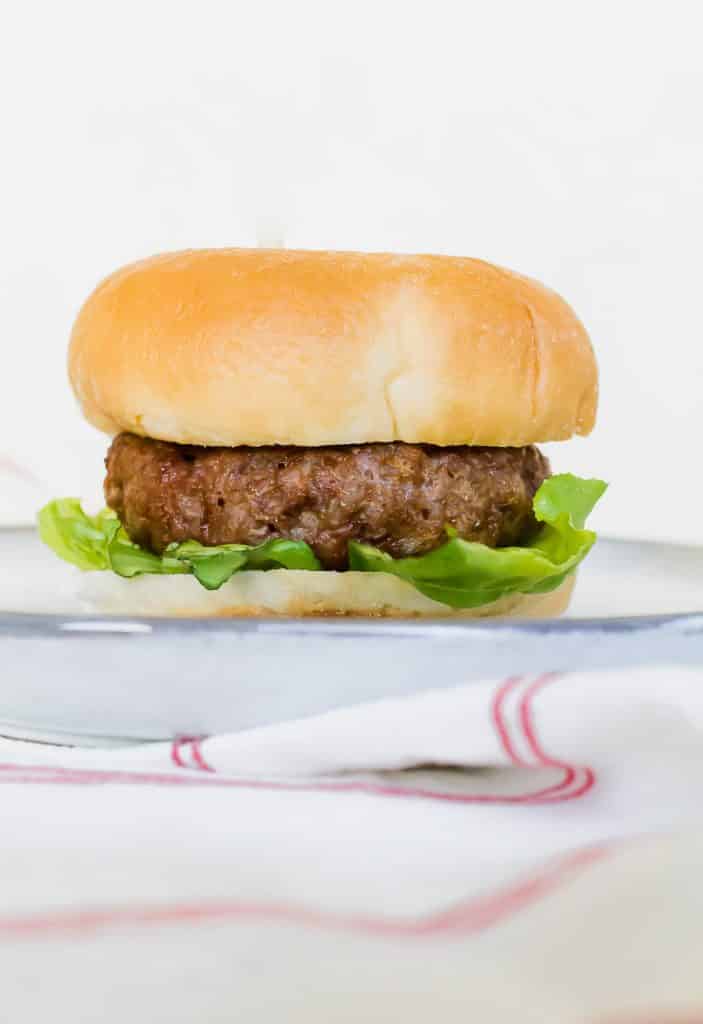 side view of an air fryer hamburger with a bun on a white plate. a red and white napkin (striped) is at the front of the vertical picture.