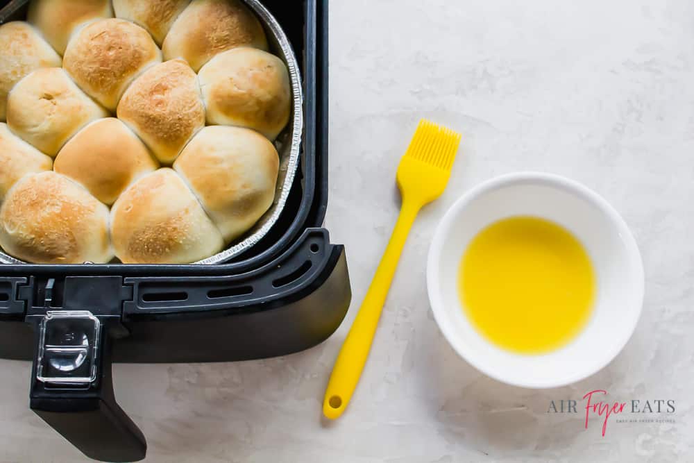 Cooked (from frozen) rolls in a black air fryer basket on the left. On the right is a white bowl with melted butter and a yellow brush