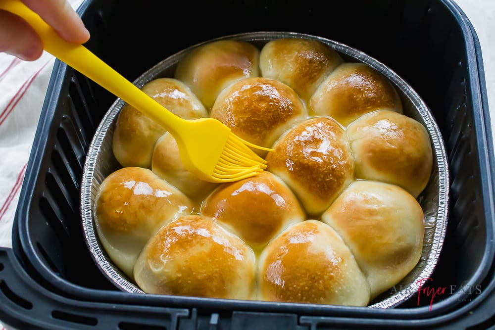Picture of air fryer frozen rolls in black air fryer basket. Yellow pastry brush is being used to spread melted butter on top of the rolls.