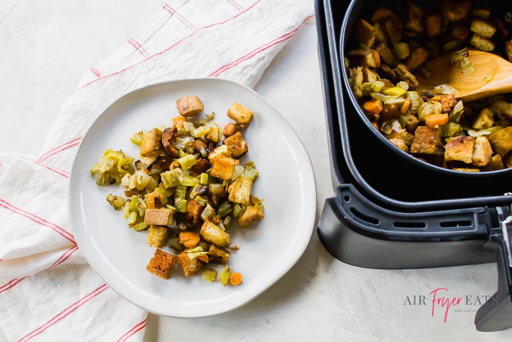 air fryer stuffing on a white plate on a white back ground with a white and red striped napkin to the left. On the right of the horizontal picture is a black air fryer basket with a pot of air fryer dressing with a wooden spoon.