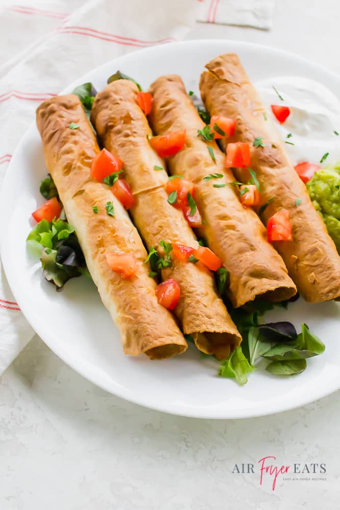 taquitos garnished with tomatoes on a white plate with a side of sour cream and guacamole