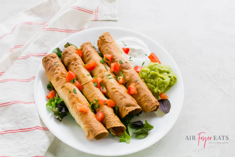 taquitos on a bed of mixed greens topped with tomatoes and garnished with guacamole and sour cream