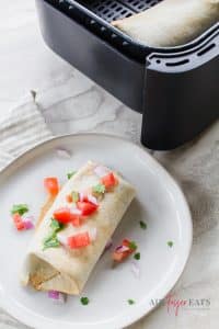 a burrito garnished with diced tomatoes and green herbs on a white plate beside a black air fryer basket