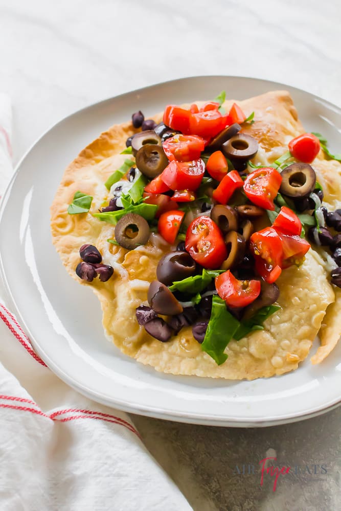 tostada topped with olives, tomatoes, black beans, and lettuce on a white plate