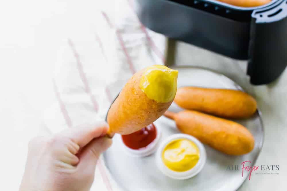 picture of two air fryer corn dogs on a white plate with ketchup and mustard. Then a left hand holding a corn dog that has been dipped into mustard