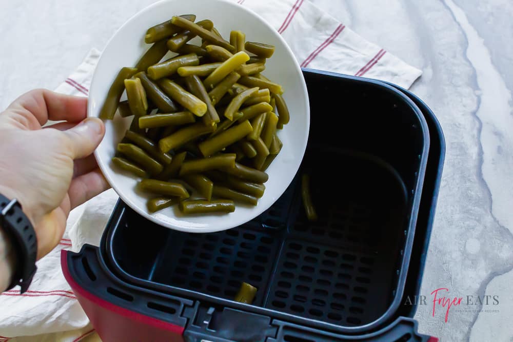 White bowl of green beans being poured into a black and red air fryer basket.