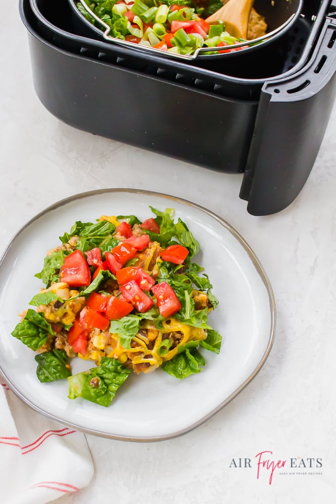 Taco dish with tomato and lettuce on a white plate next to a black air fryer basket.  