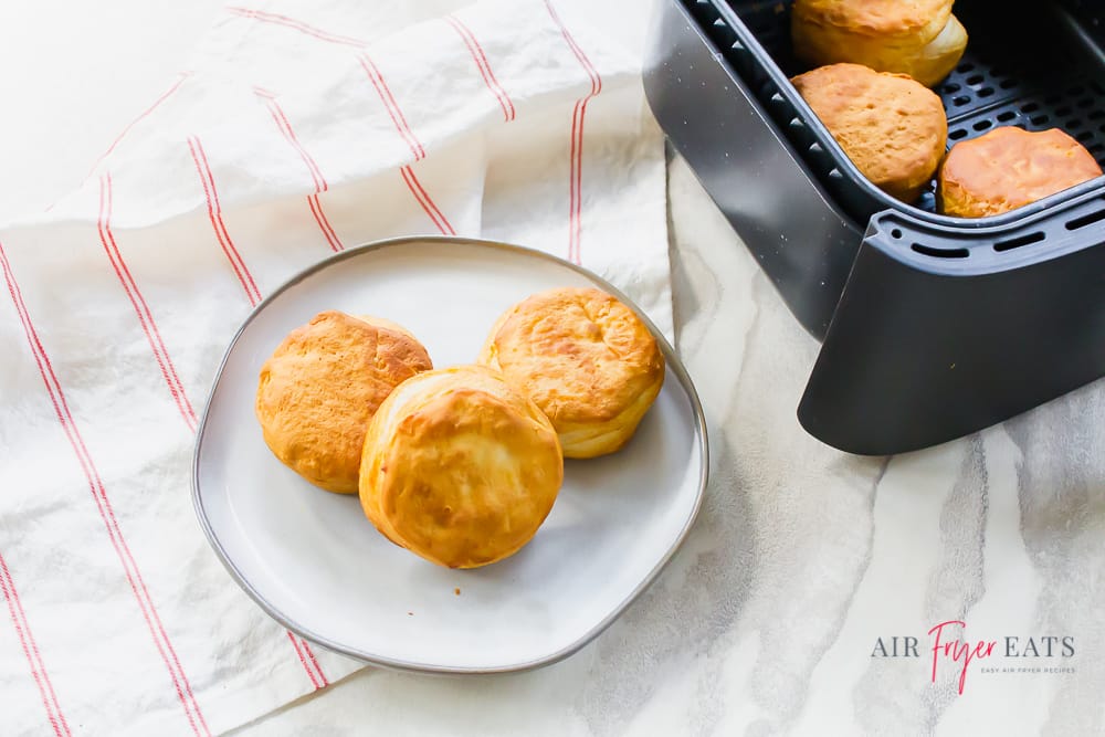 air fryer biscuits from a tub in a black air fryer basket. Basket is to the right and a white plate with biscuits is on the left.
