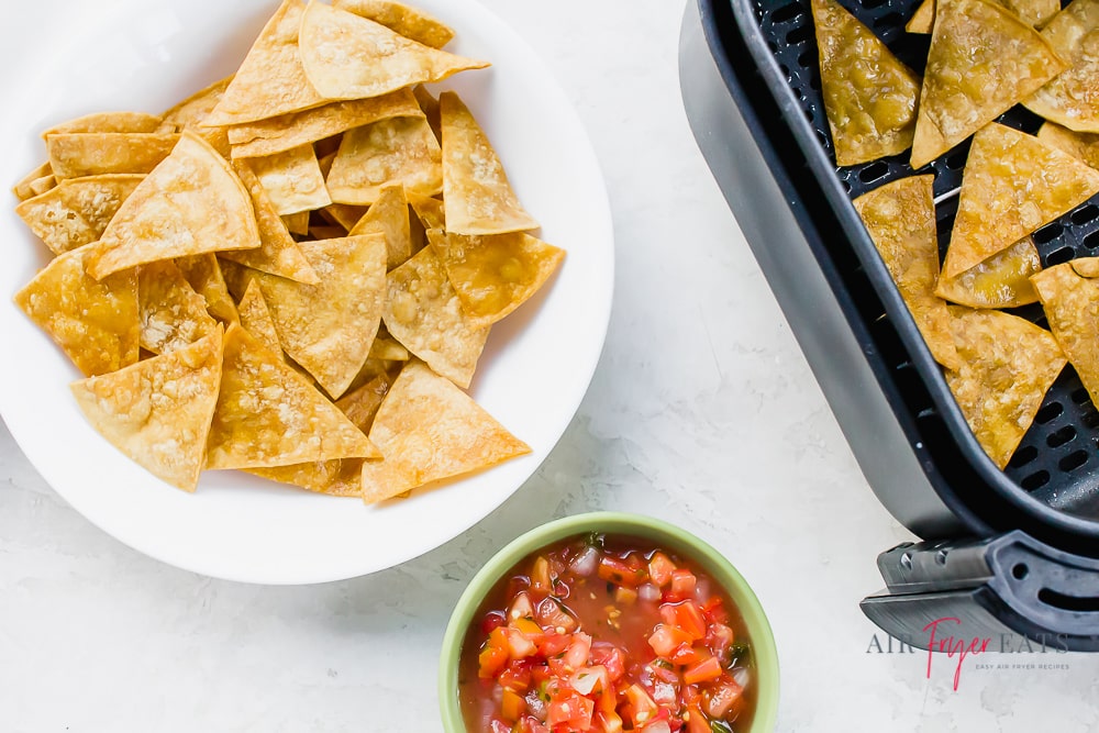 a white plate of chips with a small bowl of tomato salsa and an air fryer basket of tortilla chips