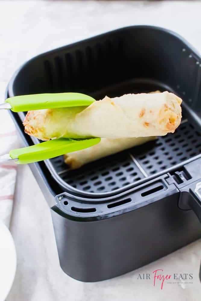 An egg roll being lifted out of an air fryer with green tongs.
