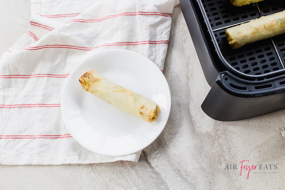 a white plate with an eggroll on it, next to an air fryer basket.