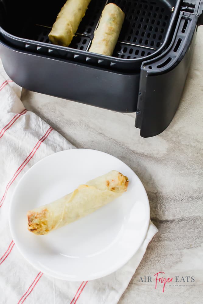 a white plate with one eggroll on it, next to an air fryer basket.