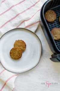 two sausage patties on a white plate next to a black air fryer basket full of sausage.