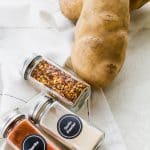 Two russet potatoes, a jar of red pepper flakes, a jar of garlic powder, and a jar of paprika, lying on a counter.
