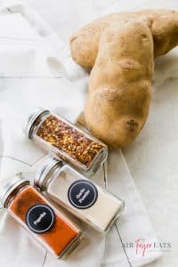 Two large potatoes are placed next to three small glass containers of red pepper flakes, paprika, and garlic powder.