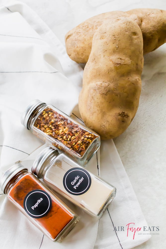 Two russet potatoes, a jar of red pepper flakes, a jar of garlic powder, and a jar of paprika, lying on a counter.