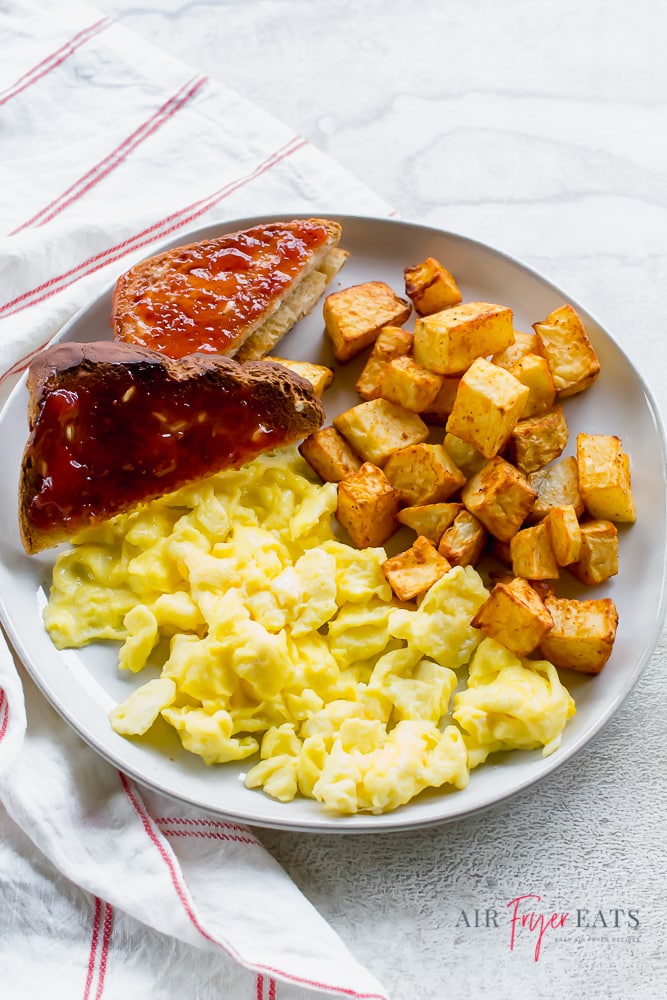 Breakfast plate that includes toast with jam, scrambled eggs, and cubed breakfast potatoes.