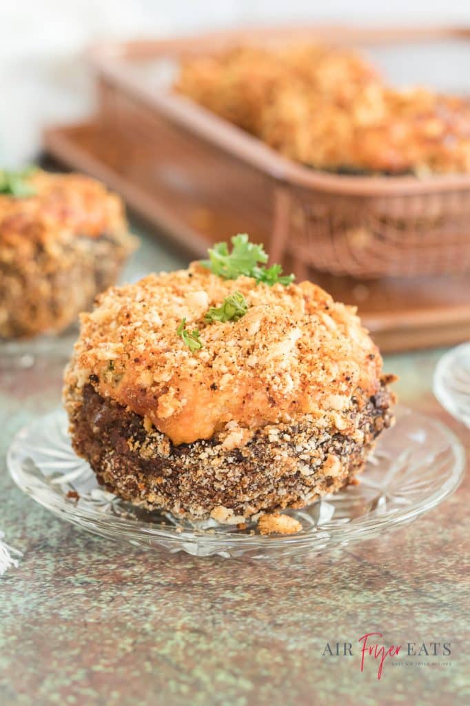 vertical picture of a stuffed and breaded mushroom on a clear glass plate and a tray of more stuffed mushrooms in the back but blurred out.