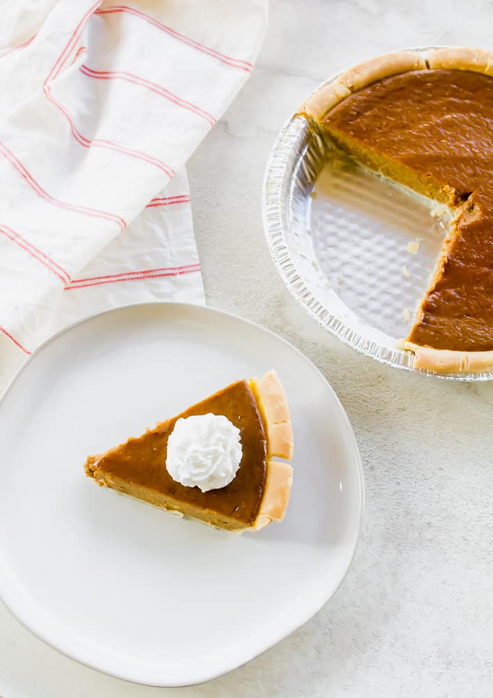Vertical picture of air fryer pumpkin pie. One slice with whipped cream is on a white plate on the bottom of the picture. The rest of the pie in a silver pie tin is at the top of the vertical photo.
