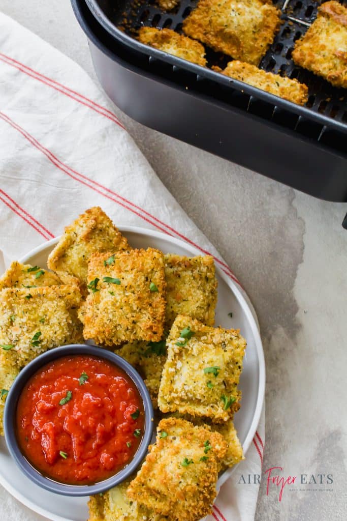 a plate of fried ravioli with a cup of marinara sauce next to an air fryer basket.