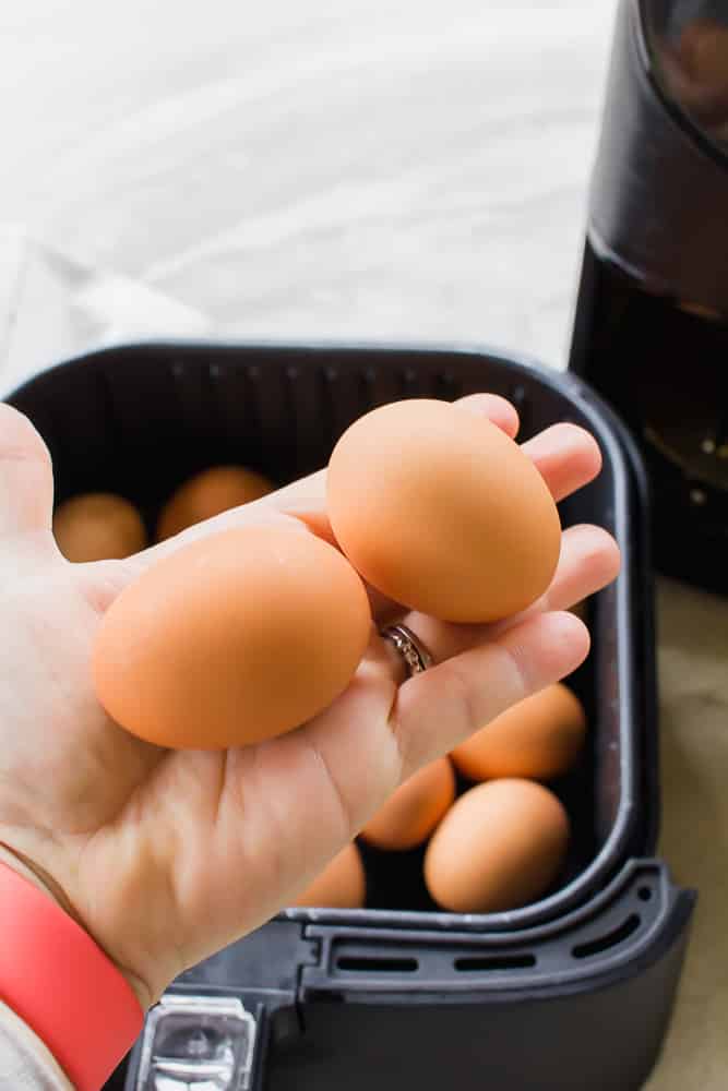 a hand holding two brown eggs over an air fryer basket full of eggs.