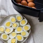 a plate of halved hard boiled eggs next to an air fryer basket full of whole eggs.