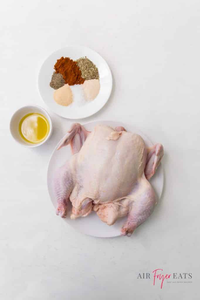 A whole chicken next to a small bowl of oil and a plate of dry rub spices