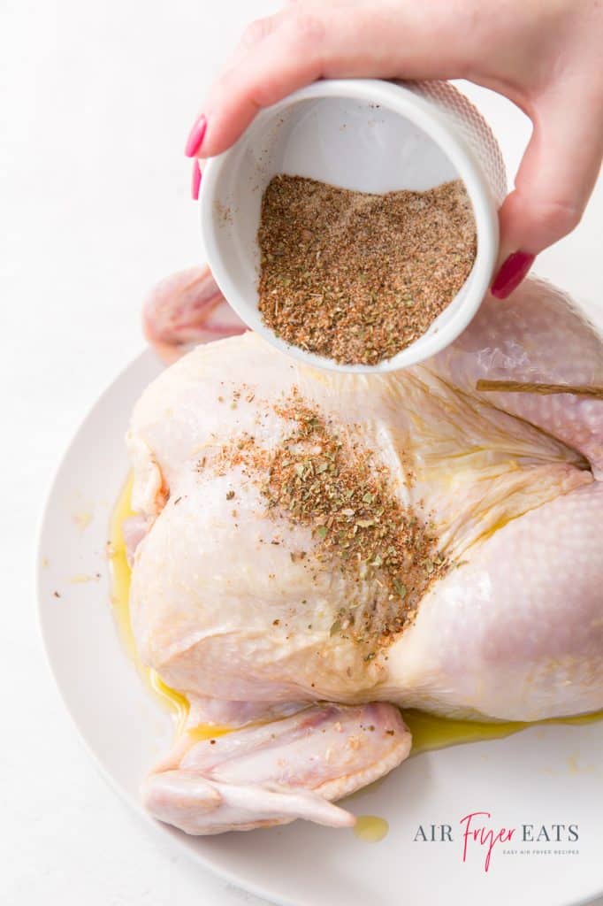 A small bowl of dry rub seasoning being poured over a whole raw chicken