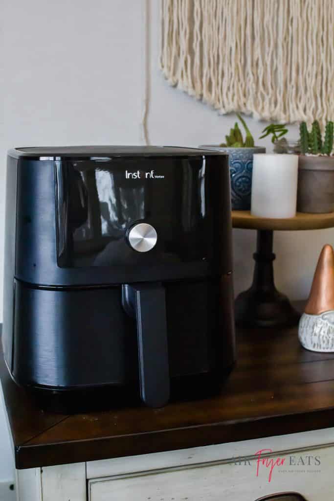 instant vortex black air fryer sitting on a buffet with a wooden top. a ghome with a copper hat sits to the right. Then a white candel with 3 catcus plants sits on a wooden top cake stand.