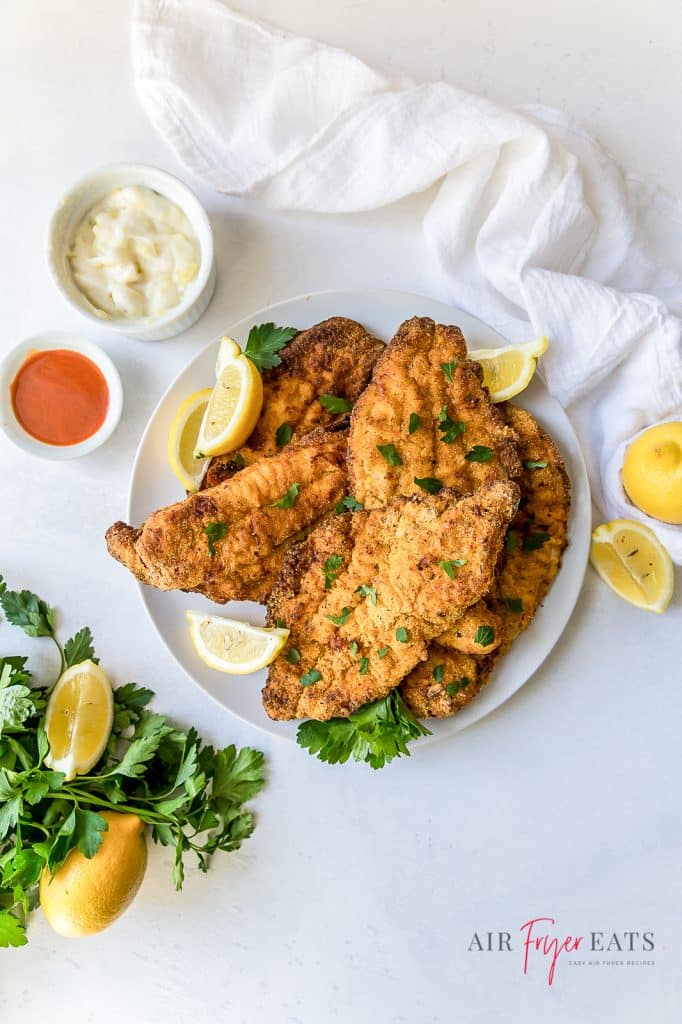 Overhead shot of a plate of fried catfish with parsley and lemon wedges next to tartar sauce and hot sauce