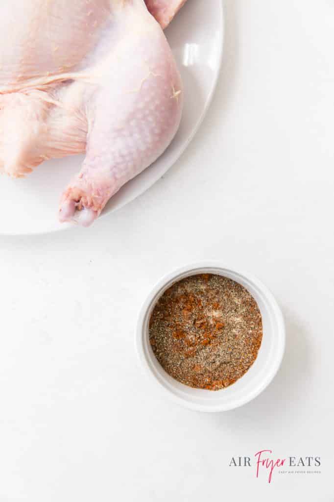 A white ramekin of dry rub seasoning next to a raw whole chicken on a white plate