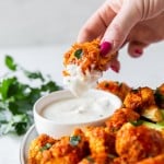 a plate of buffalo cauliflower with a cup of ranch dressing. a hand is dipping one floret into the dip.