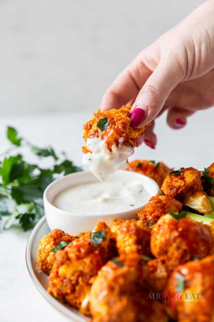 a plate of buffalo cauliflower with a cup of ranch dressing. a hand is dipping one floret into the dip. 
