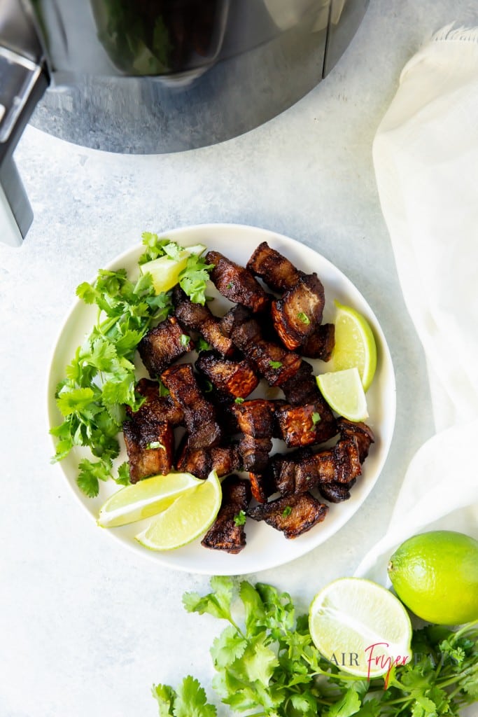 a plate of crispy pork belly garnished with cilantro and limes, next to an air fryer