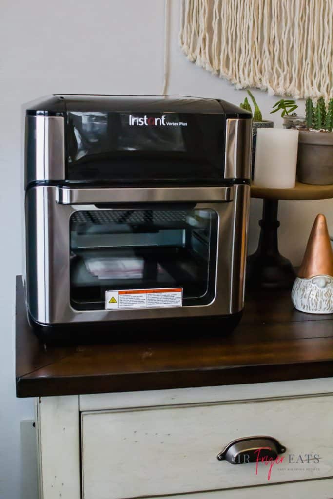 instant vortex plus air fryer oven sitting on a white buffet cabinet with a white nome with a cooper hat to the right and green catus to the top right.