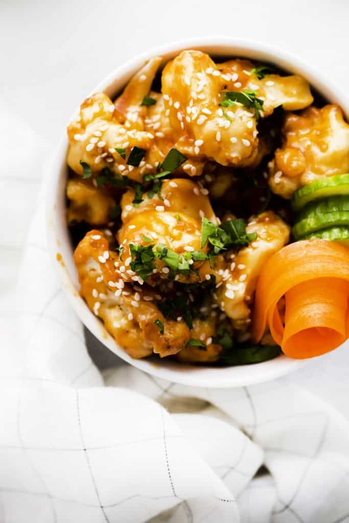 a white bowl filled with sesame cauliflower, garnished with carrot curls and fresh herbs.