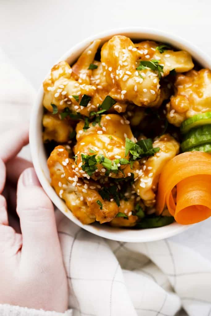 a white bowl filled with sesame cauliflower, garnished with carrot curls and fresh herbs.