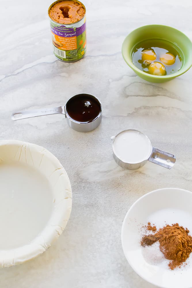 Ingredients for making pumpkin pie on a marble counter. There is a bowl of cracked eggs, a cup of maple syrup, a cup of cocunut milk, spices, a can of pumpkin and a pre-made pie shell.