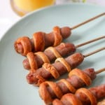 five breakfast sausages, each wrapped in strips of dough and on bamboo skewers, on a light blue/green plate.