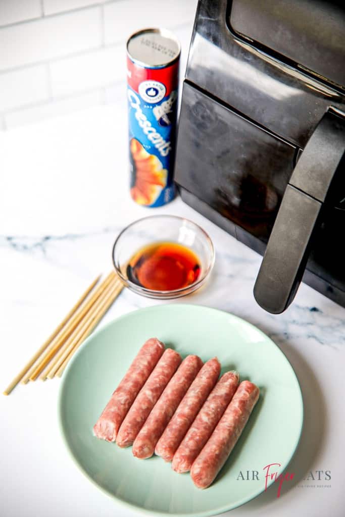 a can of crescent rools, a bowl of maple syrup, a plate of sausages and a stack of wooden skewers on a countertop next a black air fryer.