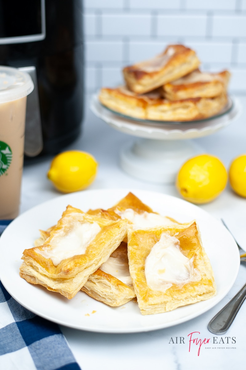 Vertical photo of white counter and back splash. Black air fryer in back left corner. Left side of photo has creamy brown liquid in plastic starbucks cup.  White plate with crispy brown dough with white cream cheese on it. Three yellowlemons in middle of picture