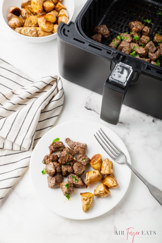 a plate of steak bites and potatoes, next to an air fryer basket